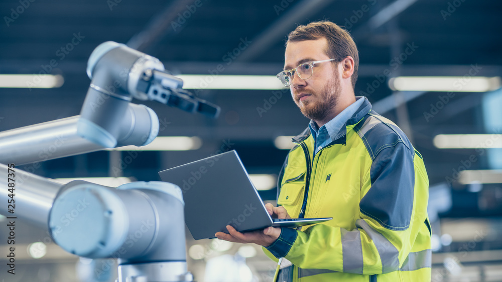 At the Factory: Automation Engineer Uses Laptop for Programming Robotic Arm. New Era in Automatic Manufacturing Industry. At the Factory: Automation Engineer Uses Laptop for Programming Robotic Arm. New Era in Automatic Manufacturing Industry.