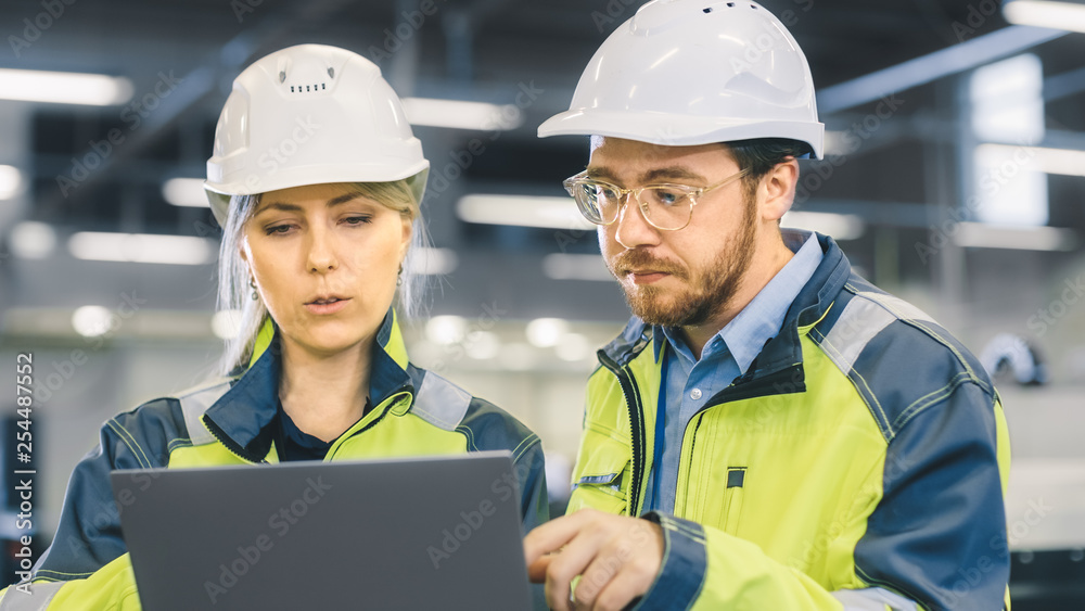 Male and Female Industrial Engineers Work on a Manufacturing Plant, They Discuss Project, Point in the Direction of the Machinery while Using Laptop. Male and Female Industrial Engineers Work on a Manufacturing Plant, They Discuss Project, Point in the Direction of the Machinery while Using Laptop.