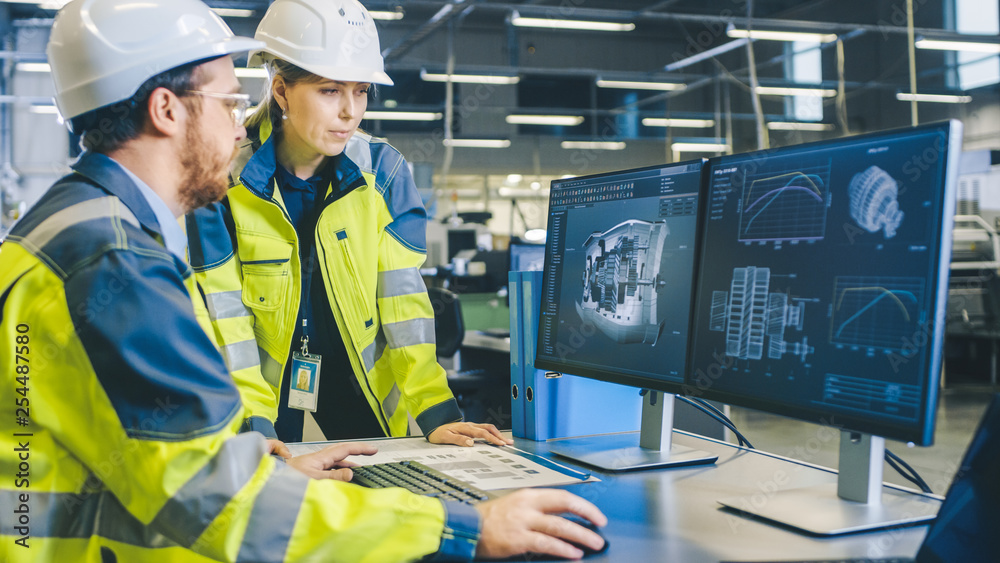 At the Factory: Male Mechanical Engineer and Female Chief Engineer Work Together on the Personal Computer, They Discuss Details of the 3D Engine Model Design. At the Factory: Male Mechanical Engineer and Female Chief Engineer Work Together on the Personal Computer, They Discuss Details of the 3D Engine Model Design.