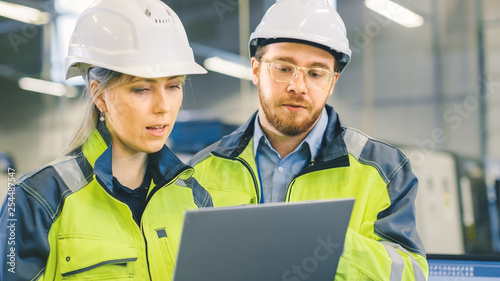 Male and Female Industrial Engineers Work on a Manufacturing Plant, They Discuss Project, Point in the Direction of the Machinery while Using Laptop. 
