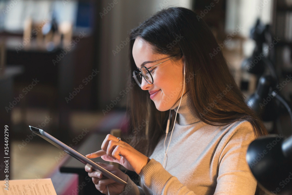 Smiling brunette college girl in earphones and glasses typing at the tablet