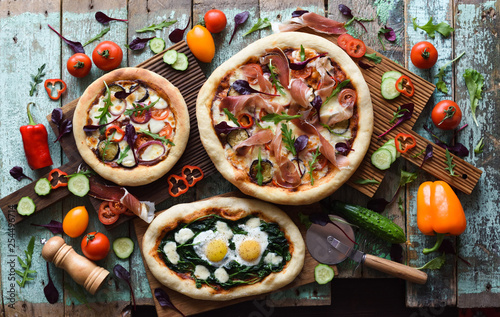 Homemade pizza party. Flatlay of puffy rustic pizzas with pancetta, aubergines, tomatoes, spinach, eggs, bell peppers and arugula served with raw vegetables and herbs on shabby blue background