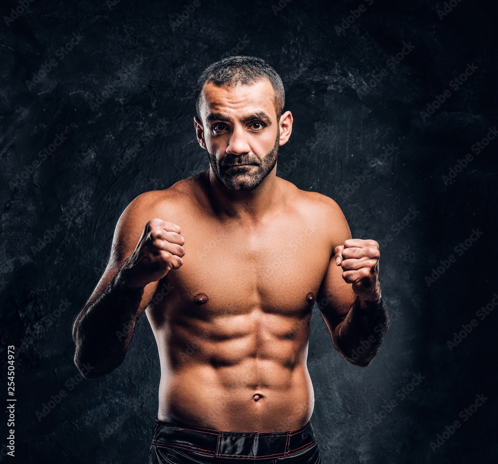 Professional Muay Thai boxer with naked torso posing for a camera. Studio photo against a dark textured wall
