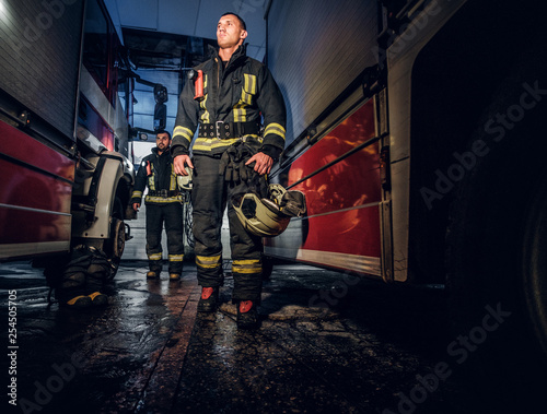 Full-length portrait of two brave firemen in protective uniform walking between two fire engines in the garage of the fire station