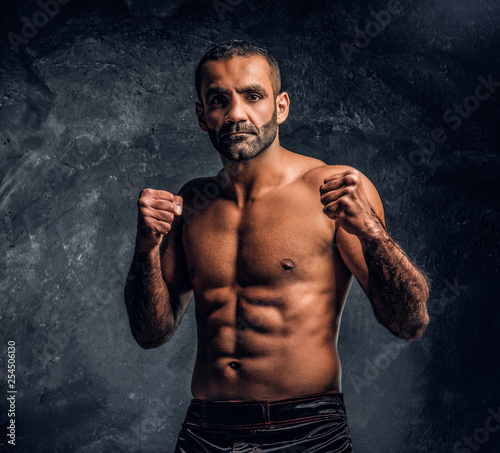 Portrait of a brutal professional fighter with naked torso posing for a camera. Studio photo against a dark textured wall © Fxquadro