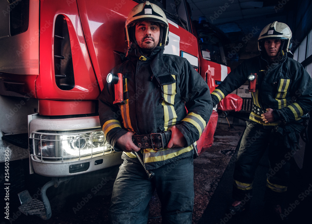 Obraz premium Dwóch strażaków w mundurach ochronnych stojących obok wozu strażackiego w garażu straży pożarnej.