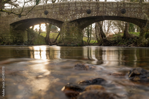 The River Teign glides under an Ancient stone bridge just outside Chagford in Devon, England, out of focus rocks in the foreground pointing towards the bridge