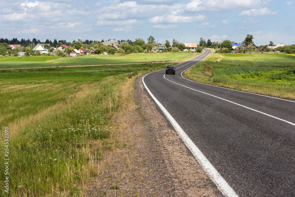 View of the small asphalt road in the countryside of Belarus with blue sky and cumulus clouds on the background.