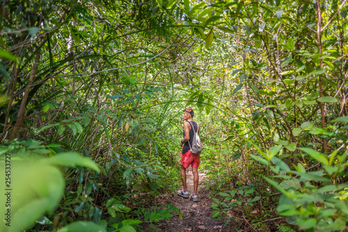 Young man walks along the path through bush and tree thickets wearing shorts, top tank, sneakers and a light backpack. A hiker in a national park strolls through lush vegetation.