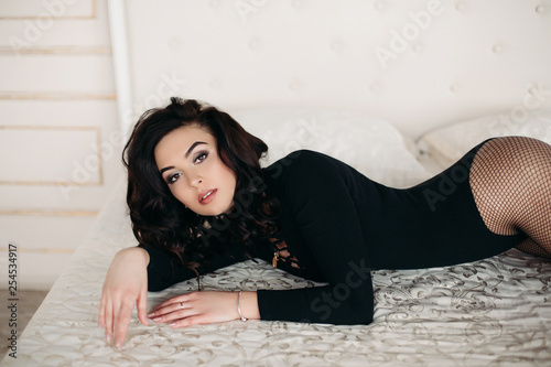 Front view of beautiful and sexy woman in black lying on big bed, looking at camera, holding legs up. Attractive lady with plups lips and posing, looking at camera. Passion and beauty. photo