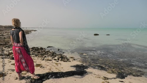 Caucasian woman looking turquoise clear sea at Persian Gulf near Al Areesh Fishermens villages, Northerm of Qatar. Happy tourist visits abandoned village, Middle East, Arabian Peninsula.Sunny blue sky photo