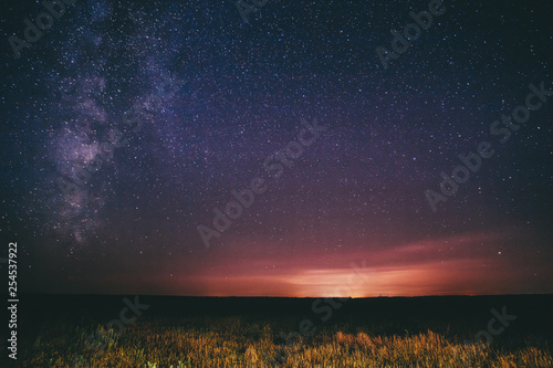 Night Starry Sky Above Field And Yellow City Lights On Background. Night View Of Natural Glowing Stars