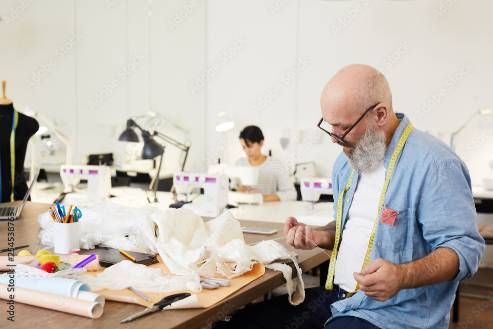 Obraz premium Senior man in shirt and eyeglasses preparing white threads for sewing one of items of new fashion collection while working in tailoring shop