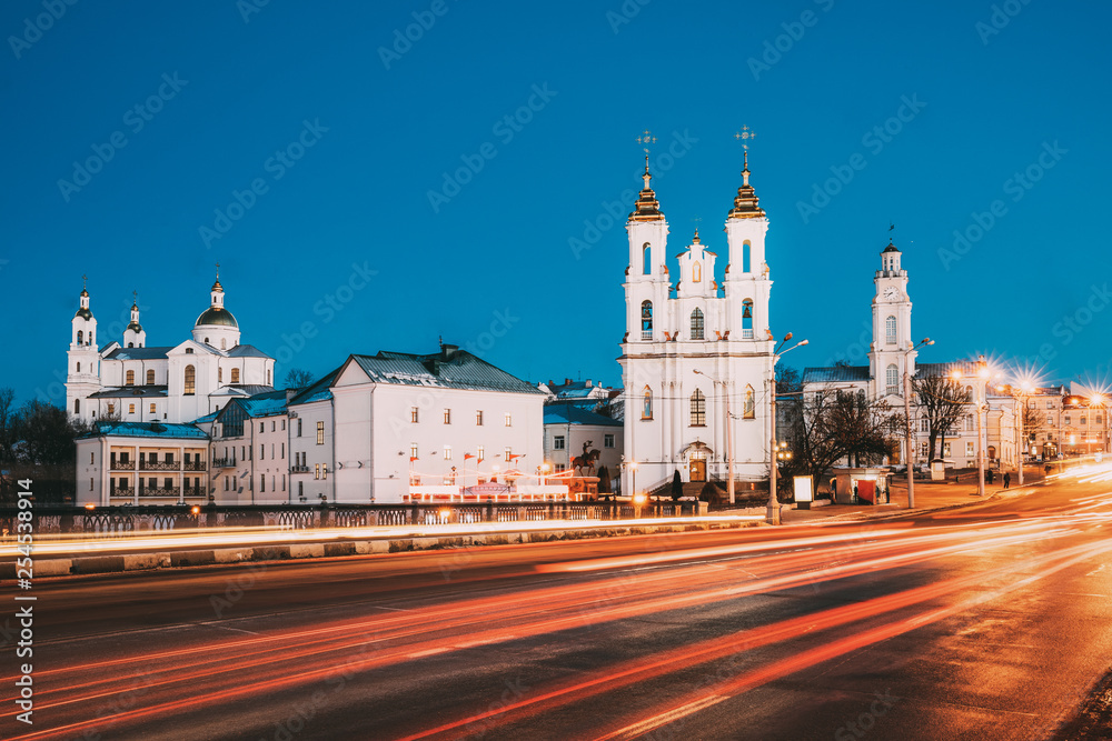 Naklejka premium Vitebsk, Belarus. Traffic At Street And Holy Assumption Cathedral, Holy Resurrection Church And City Hall In Evening Or Night Illumination At Winter