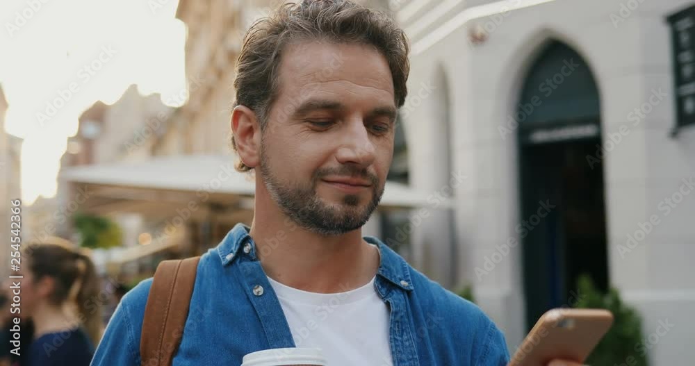 Caucasian handsome man with coffee to go walking on the street and texting on the smartphone. Close up.