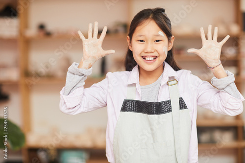 Waist up portrait of happy Asian girl posing in pottery workshop showing hands covered with clay to camera, copy space