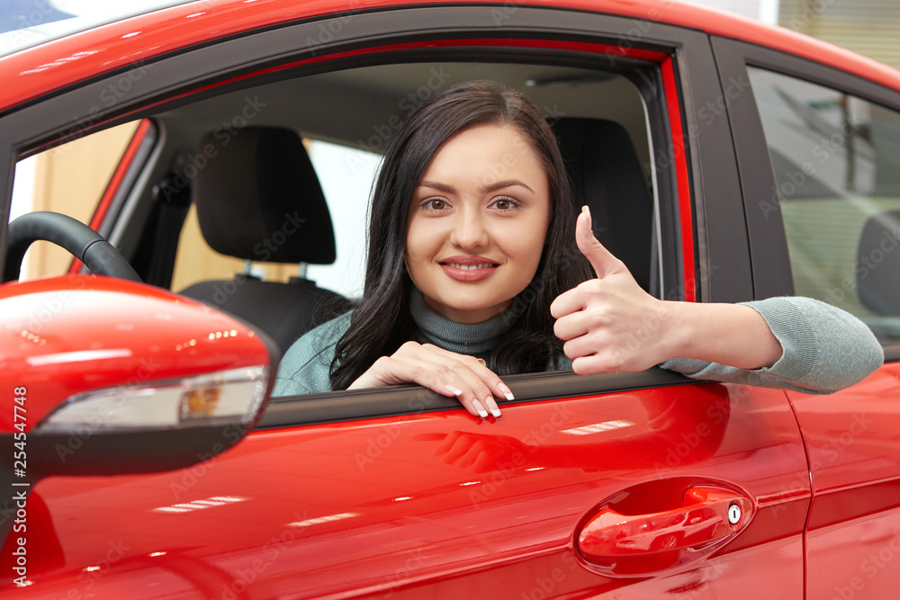 Her first car. Portrait of a beautiful cheerful brunette female smiling to the camera showing thumbs up posing sitting in a red car at the car salon