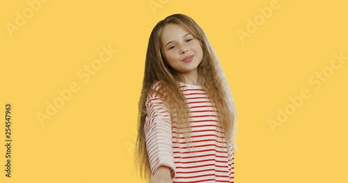 Portrait shot of the blond pretty teen girl with long hair in the striped blouse blowing kisses while standing on the orange wall background.