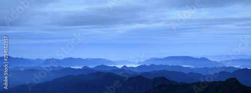 Abstract Image, Mountain Silhouettes at dawn - rolling jagged mountain peaks, cold blue color hues. Panoramic Abstract Background Image, overcast skies, layers of rolling mountains in the distance.