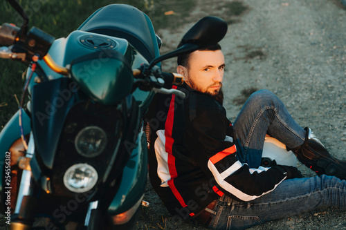 Portrait of a bearded confident man sitting in the ground leaning on his motorcycle looking away seriously while exploring new places in his journey.