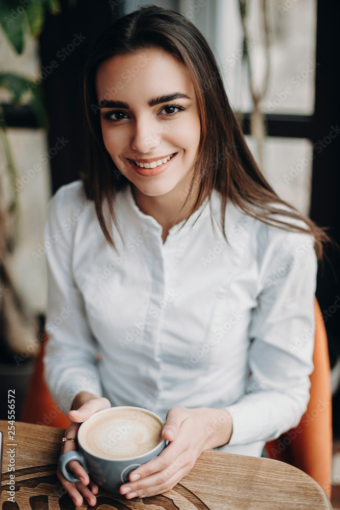 Portrait of a beautiful long hair business woman sitting in a cafe and smiling while looking at camera with a cup of coffee in her hands at lunch break. Portrait of a beautiful long hair business woman sitting in a cafe and smiling while looking at camera with a cup of coffee in her hands at lunch break.
