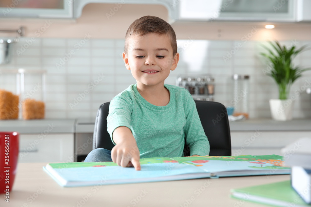 Cute little boy reading book at table in kitchen Cute little boy reading book at table in kitchen