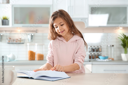 Cute little girl reading book at table in kitchen