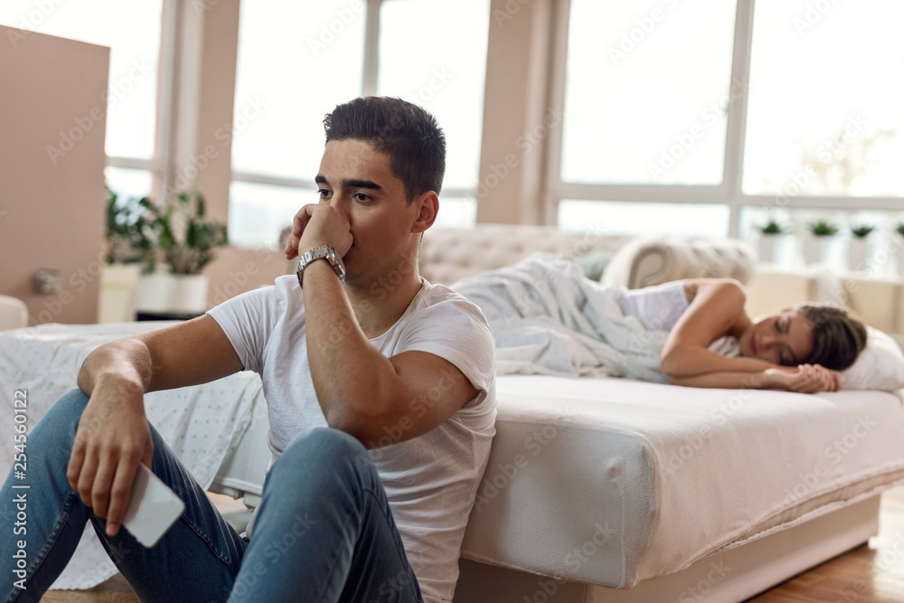 Worried man feeling sad while his girlfriend is sleeping in the background. Worried man feeling sad while his girlfriend is sleeping in the background.