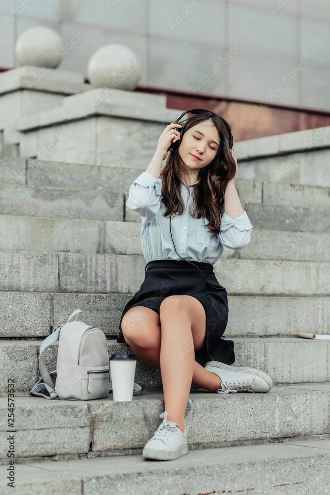 A beautiful student sits on the steps in a shirt and skirt,listening to music in headphones ,resting on the steps of the University in the fresh air. Concept of education