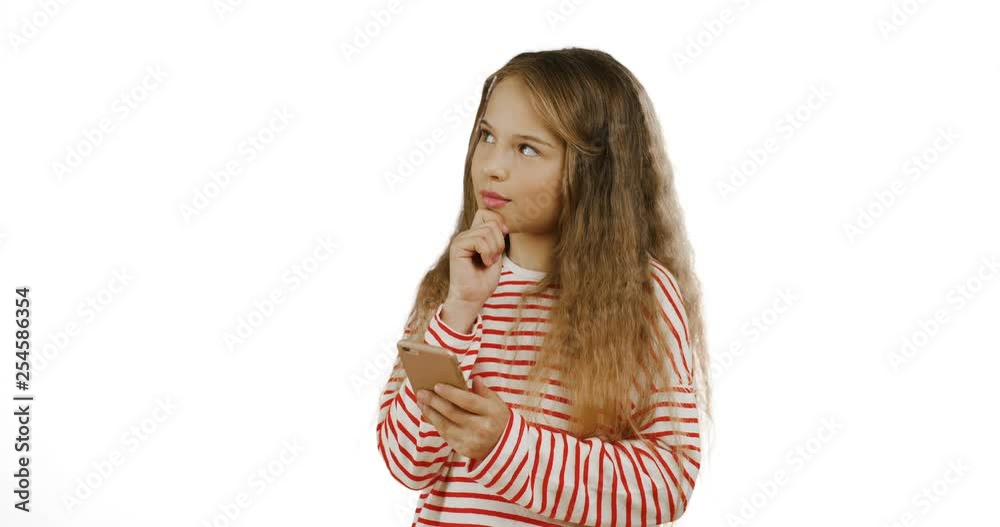 Adorable small teen girl with long fair hair standing on the white background, thinking and scrolling on the smartphone screen. Adorable small teen girl with long fair hair standing on the white background, thinking and scrolling on the smartphone screen.