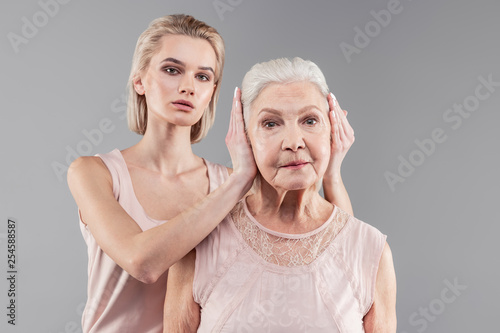 Neutral good-looking young lady tightly closing ears of old mother