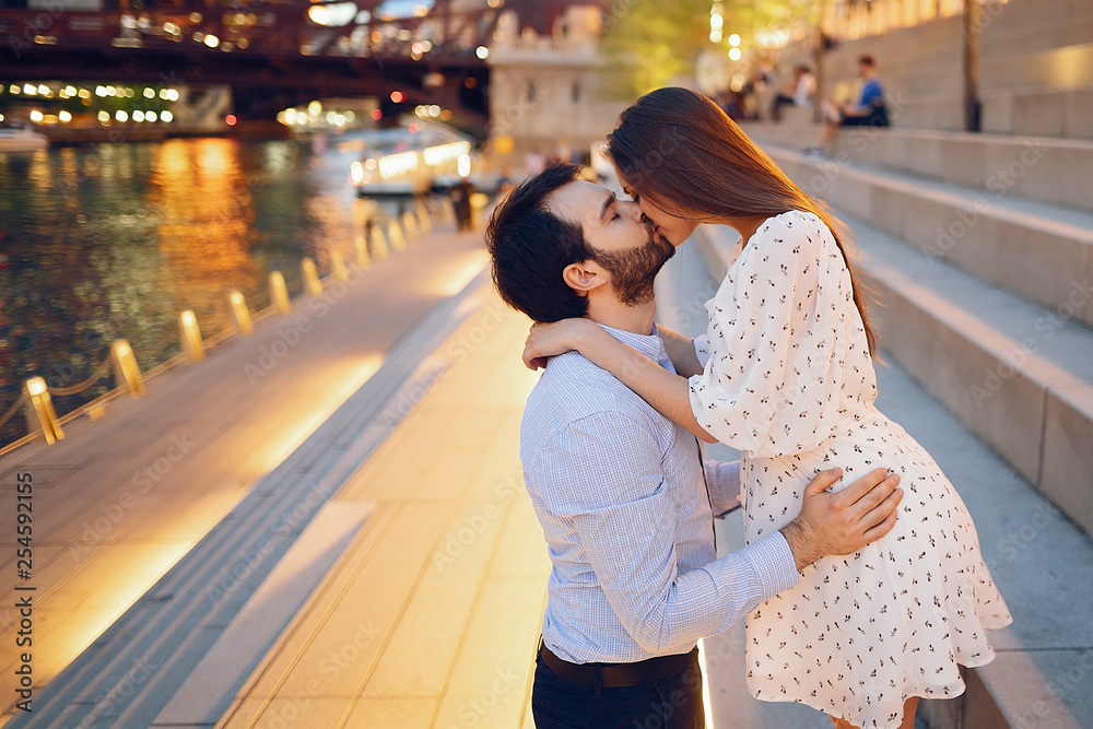 Naklejka premium beautiful long-haired girl in summer dress with her handsome husband in white shirt and pants walking in a evening Chicago