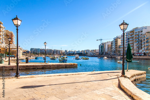 San Giljan, Malta, on January 7, 2019. Walking and fishing boats in the picturesque bay Spinola are lit with the sun in the winter morning. beautiful embankment in the distance photo