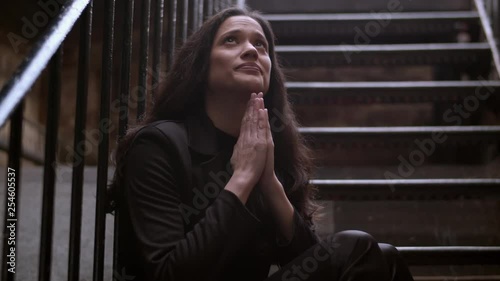 Medium shot of a beautiful mixed heritage woman sitting on some stairs outdoors and praying looking up to heaven