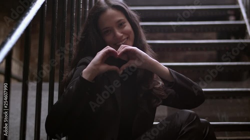 Static shot of an attractive woman making a heart symbol with her hands while sitting partially turned on some stairs outdoors with a slight simile
