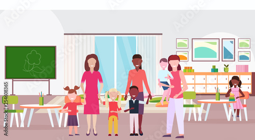 women teachers teaching mix race boys and girls preschool modern kindergarten children classroom with chalkboard desks chairs kid room interior full length flat horizontal