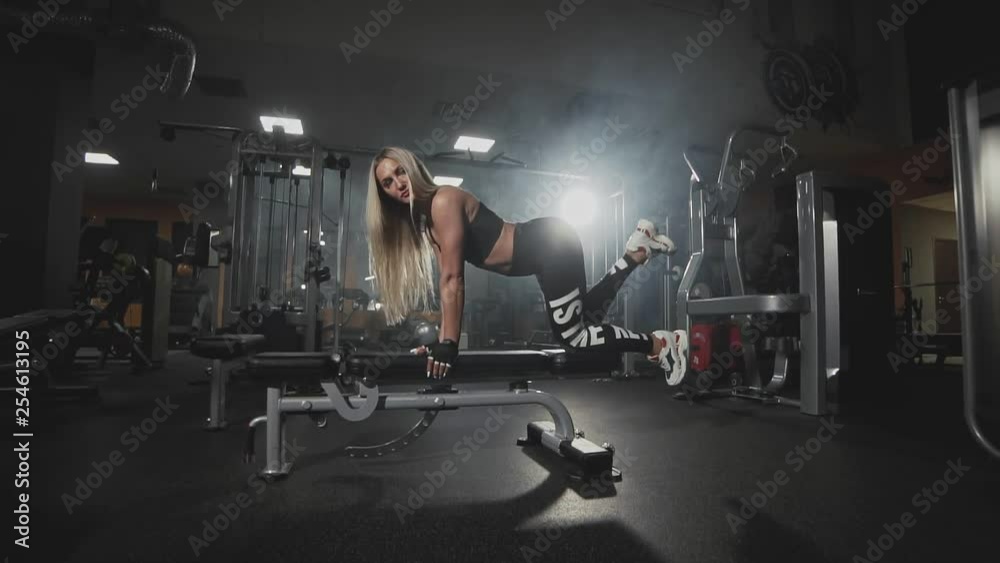 Beautiful girl doing kick backs on bench at dark smoky gym in slow motion. Sexy blonde woman training in sport center. Wide angle. Healthy lifestyle. Beautiful girl doing kick backs on bench at dark smoky gym in slow motion. Sexy blonde woman training in sport center. Wide angle. Healthy lifestyle.