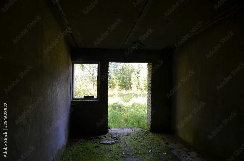View of the green field and trees from the dark destroyed old brick building with a mossy floor through the door and window View of the green field and trees from the dark destroyed old brick building with a mossy floor through the door and window