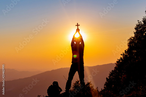 human praying to the GOD while holding a crucifix symbol with bright sunbeam on mountain at sunset