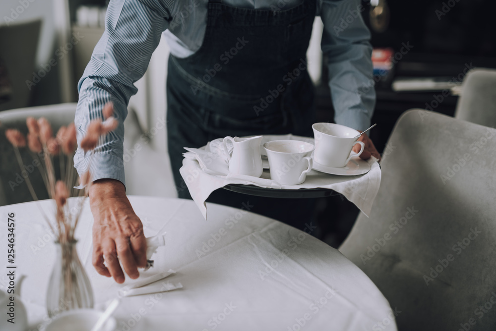 Fototapeta premium Waiter in apron holding tray with cups of coffee and placing sugar on table