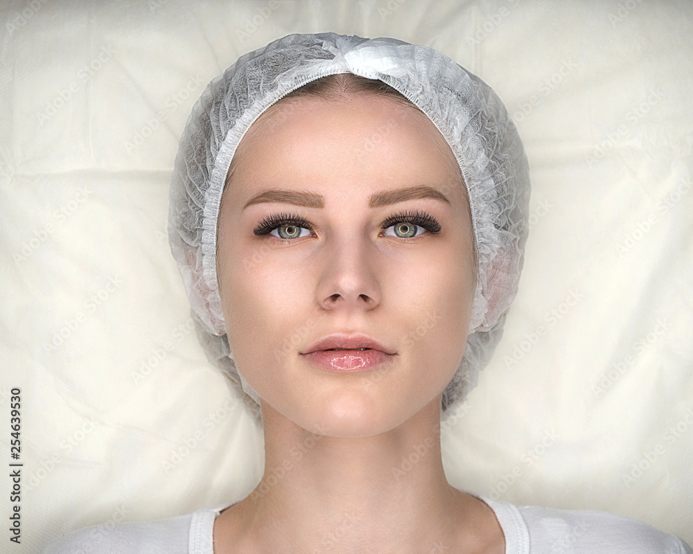 Female face with new false lashes and in disposable cap. Woman after eyelash extension procedure is lying on the couch. Close up, top view.