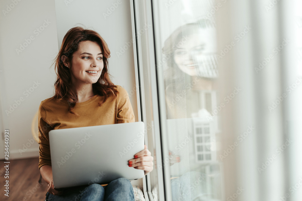 Dreamful female sitting near window with laptop