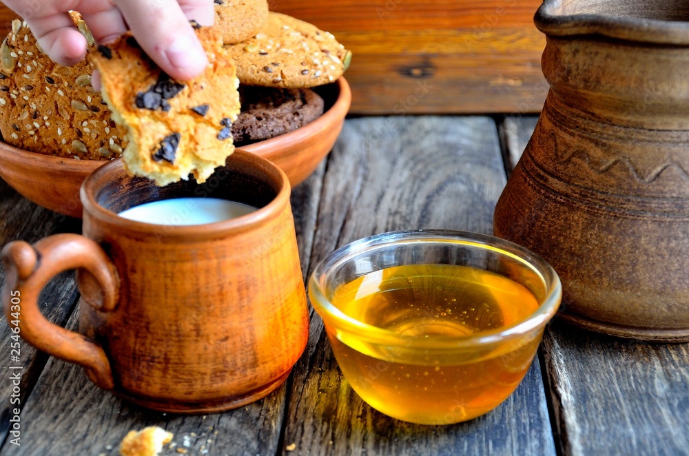 Fototapeta premium Oatmeal cookies in a ceramic rustic plate, milk in a ceramic mug, napkin, honey on a rustic wooden table.