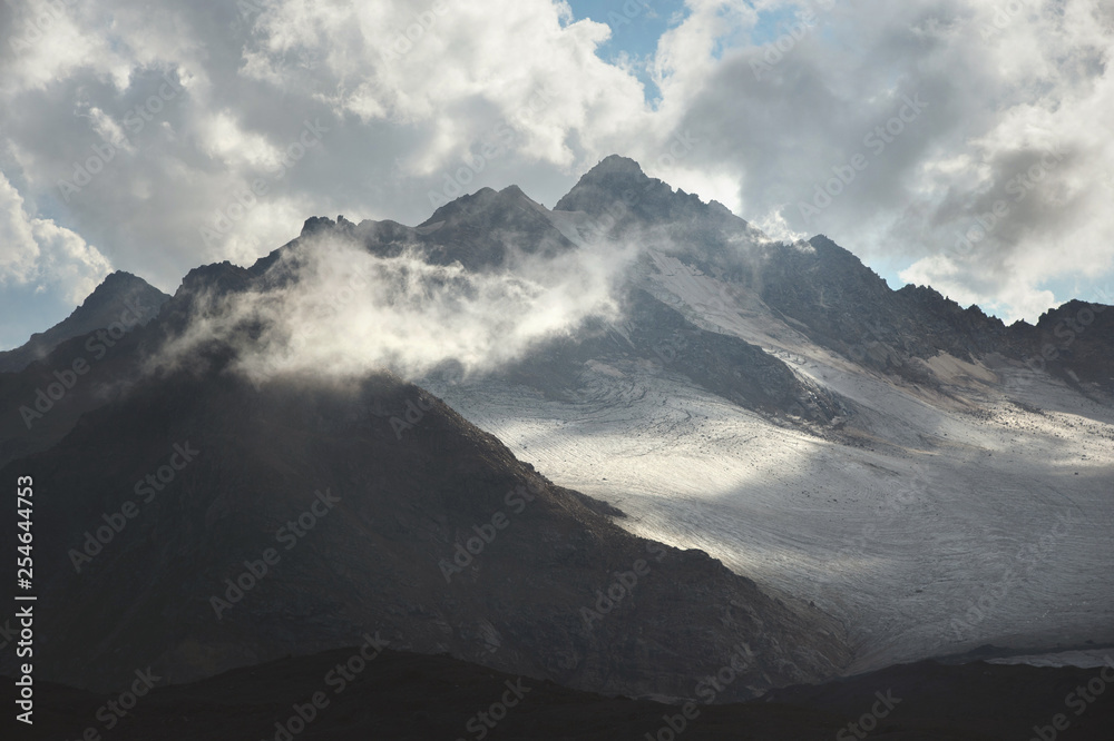Mountain landscape of a high mountain slope with a cracked glacier muddy volcanic slope against the backdrop of the Caucasus Mountains in the window of clouds. Glaciers of the North Caucasus