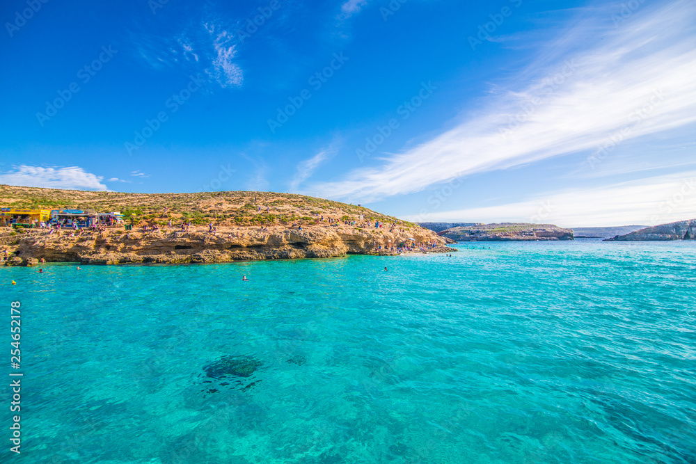 Fototapeta premium Comino, Malta - listopad 2018: Turyści tłoczą się w Błękitnej Lagunie, aby cieszyć się czystą turkusową wodą w słoneczny letni dzień z czystym błękitnym niebem i łodziami na wyspie Comino na Malcie.