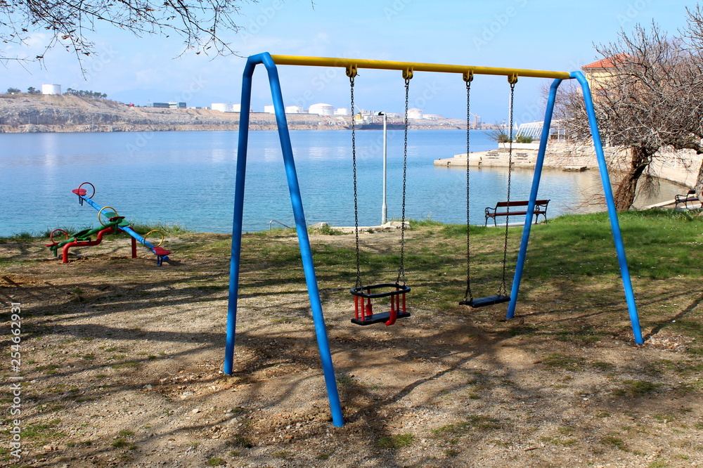 Outdoor public playground equipment in shape of swing and seesaw surrounded with grass and public bench with industrial complex and oil tanker in background on warm spring day Outdoor public playground equipment in shape of swing and seesaw surrounded with grass and public bench with industrial complex and oil tanker in background on warm spring day