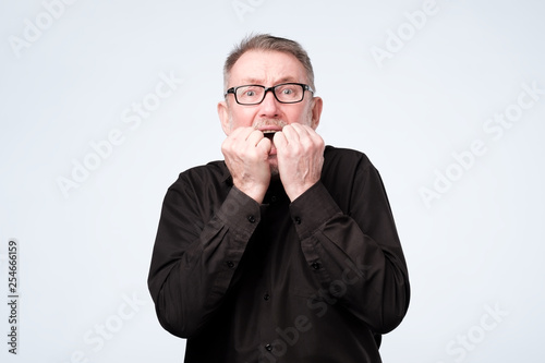 Nervous senior man in glasses bites nails, looks worried before visiting doctor