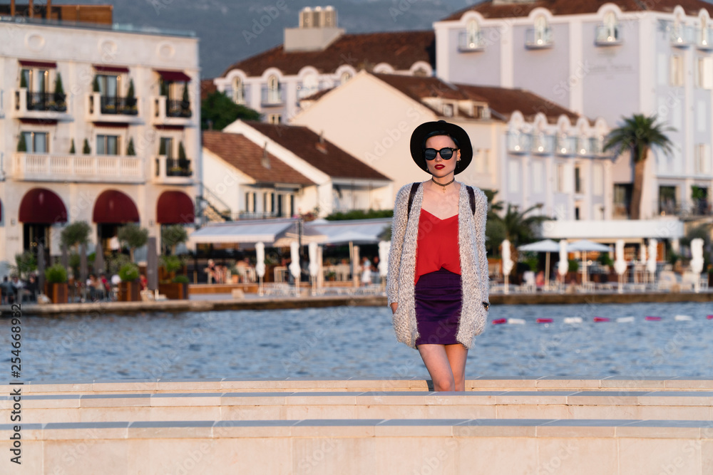 A girl in sunglasses and a black hat is standing on a pier near the sea in a European resort town on the background of luxury houses and palm trees. Travel and Vacation