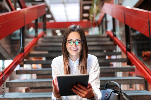 Close up of smiling female student with eyeglasses and brown hair using tablet while sitting on the stairs. Next to her bag. photo