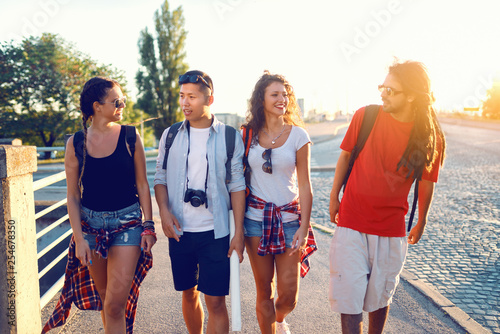 Four cheerful multicultural tourists with backpacks on backs walking down the street and chatting. Summertime vacations concept.
